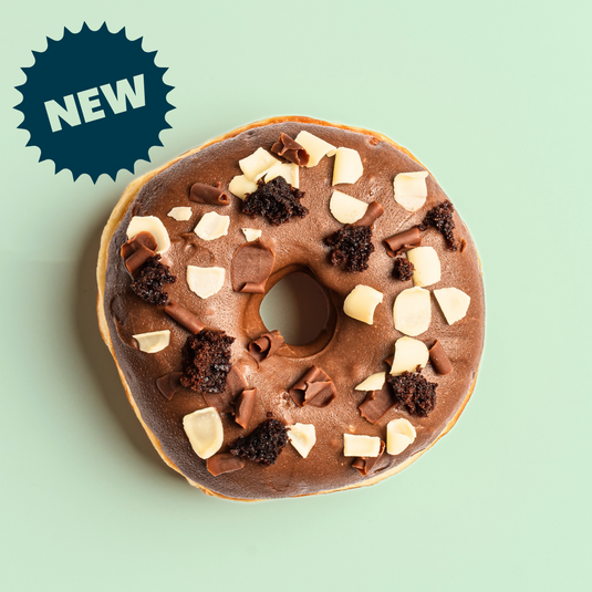 Chocolate ring doughnut with milk & white chocolate curls and brownie pieces on a light green background, featuring a 'NEW' label.